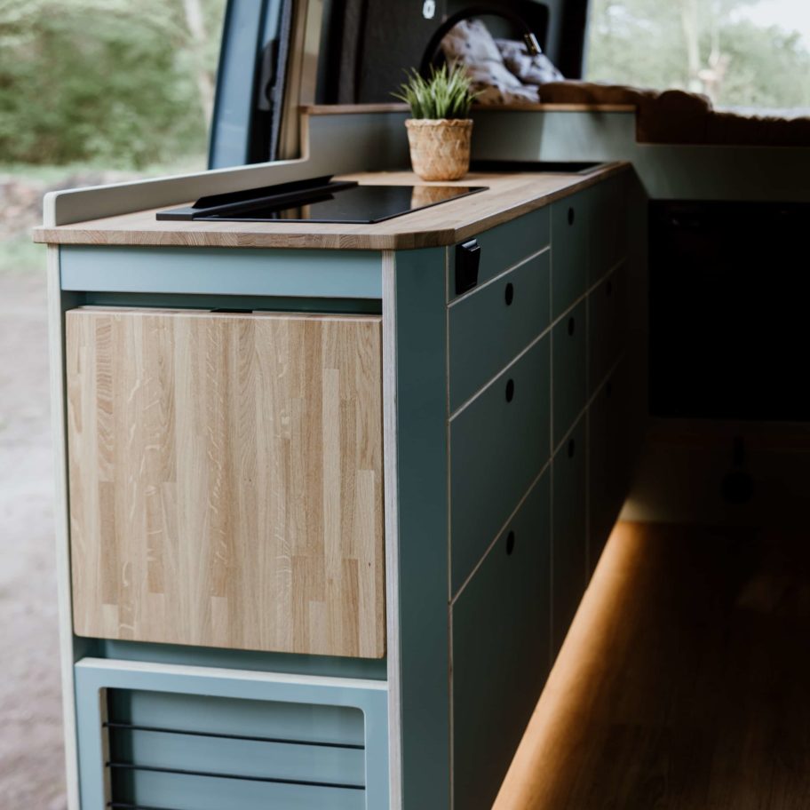 Cuisine sur mesure du Sprinter L2H2 Aoraki – mobilier bleu-gris, plan de travail bois clair et éclairage d’ambiance.