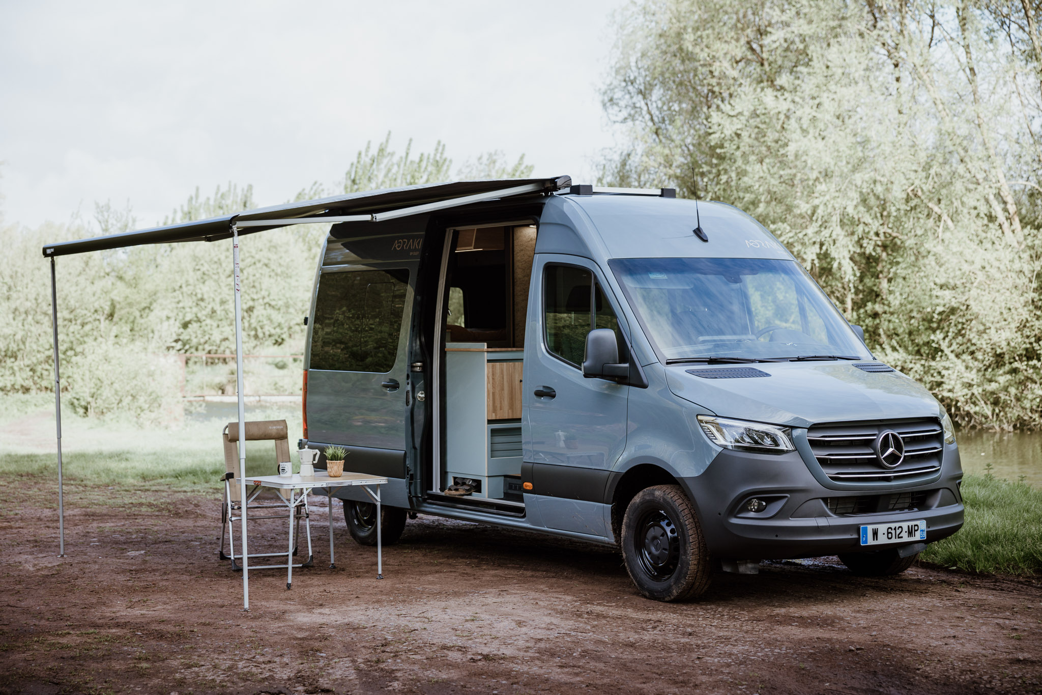 Vue 3/4 extérieure du Mercedes Sprinter L2H2 Aoraki aménagé par OLEY WORKSHOP – store Fiamma ouvert et mobilier extérieur installé, prêt pour l’aventure.