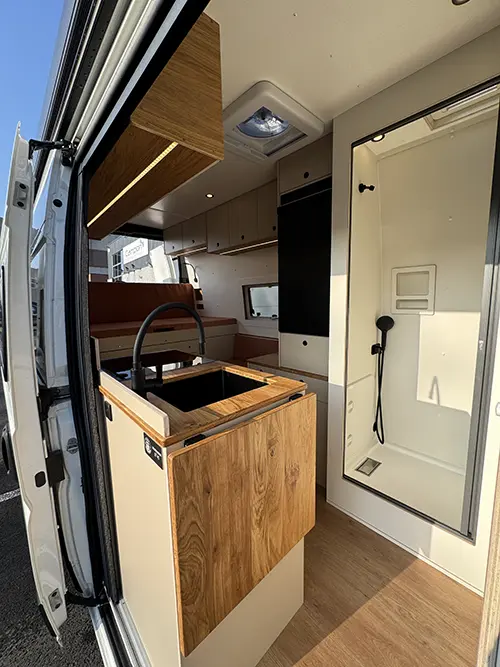 Aménagement de fourgon avec cuisine compacte en bois, douche intégrée et vue sur l’espace salon.