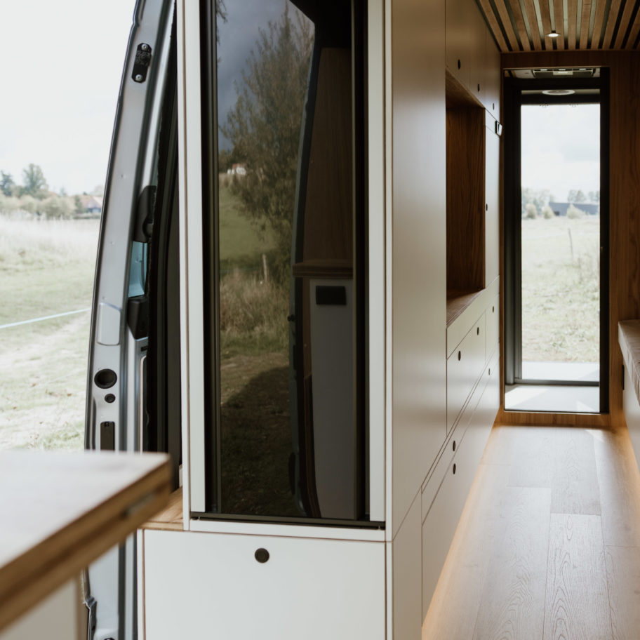 Vue latérale intérieure du fourgon avec cuisine à gauche et entrée ouverte sur l’extérieur.