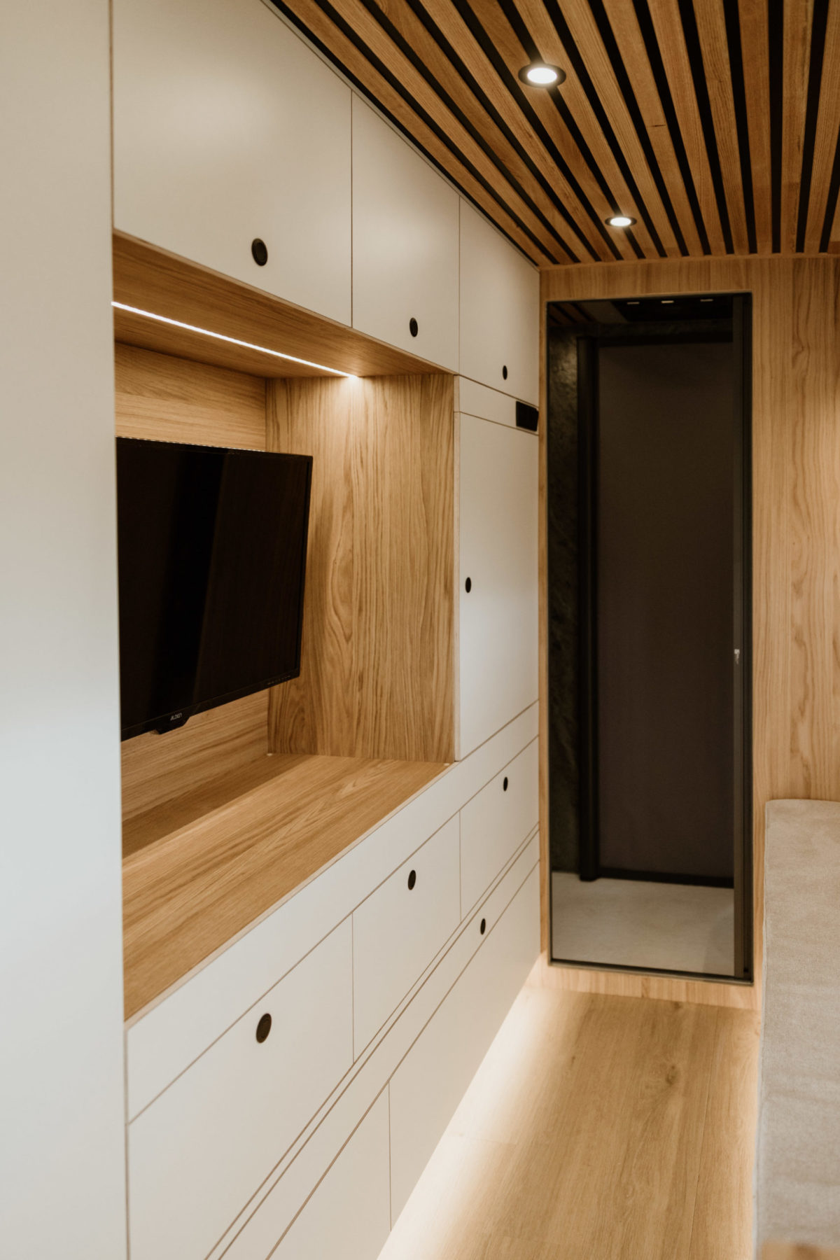 Vue intérieure du Mercedes Sprinter 4x4 L4H2 519 CDI “Signature” par OLEY WORKSHOP – meuble TV intégré, coin banquette confortable et finitions en bois clair.