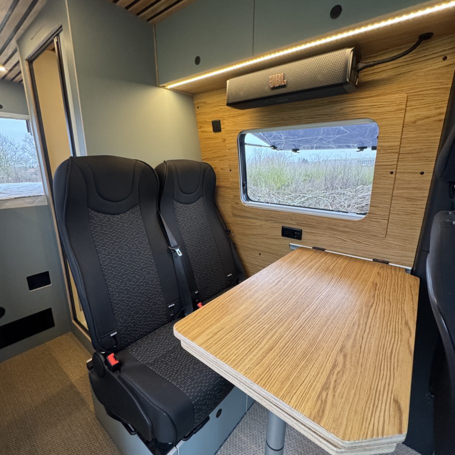Espace repas d’un van Mercedes Sprinter aménagé avec table en bois et deux sièges passagers.