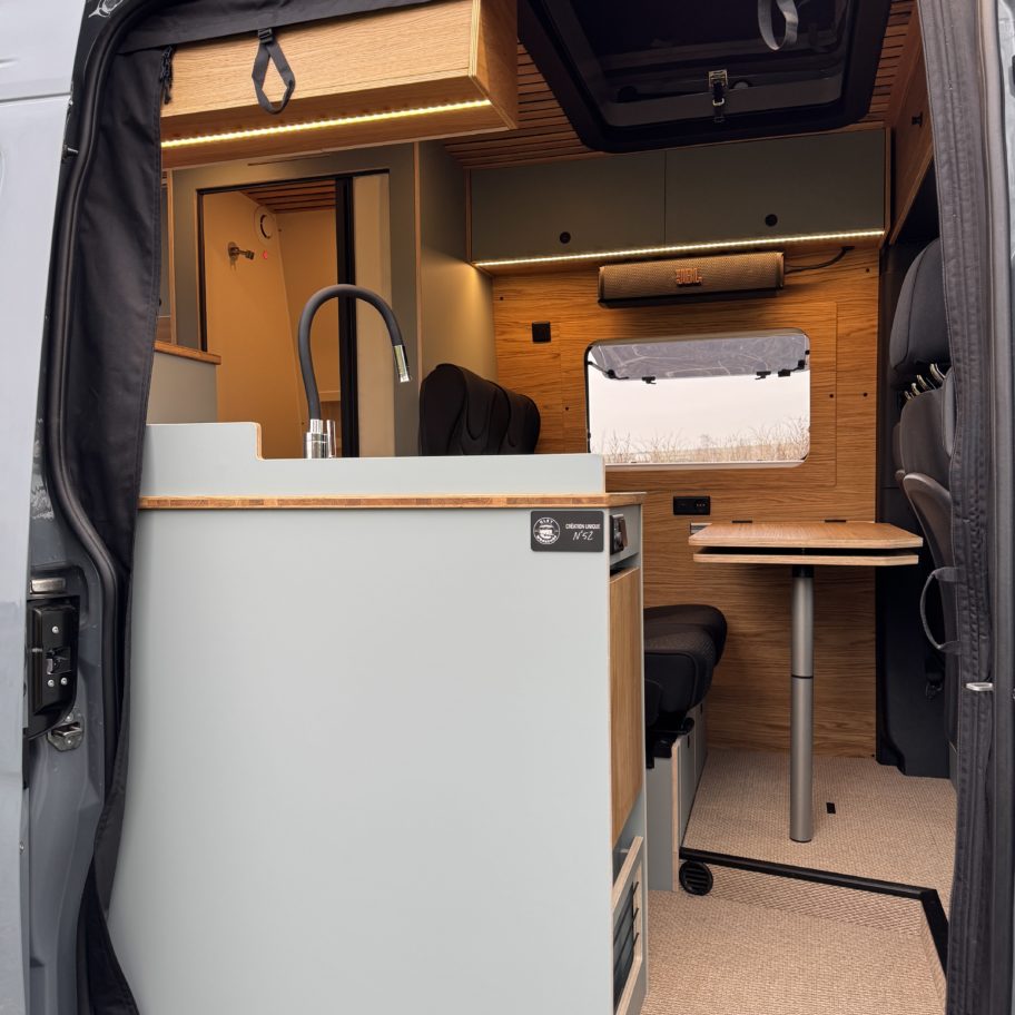 Vue arrière d’un van Mercedes Sprinter aménagé avec meuble cuisine, évier, plan de travail et rangements en bois.