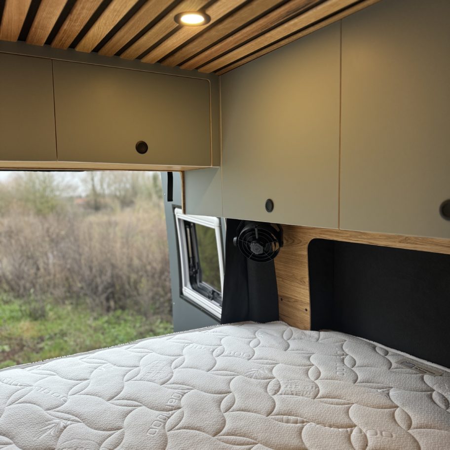 Lit double d’un van Mercedes Sprinter aménagé avec rangements hauts et fenêtre latérale.