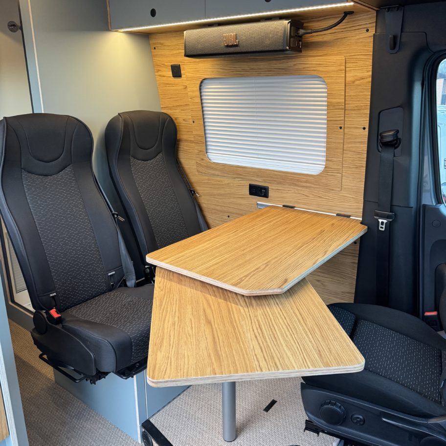 Espace repas d’un van Mercedes Sprinter aménagé avec table en bois et deux sièges passagers.