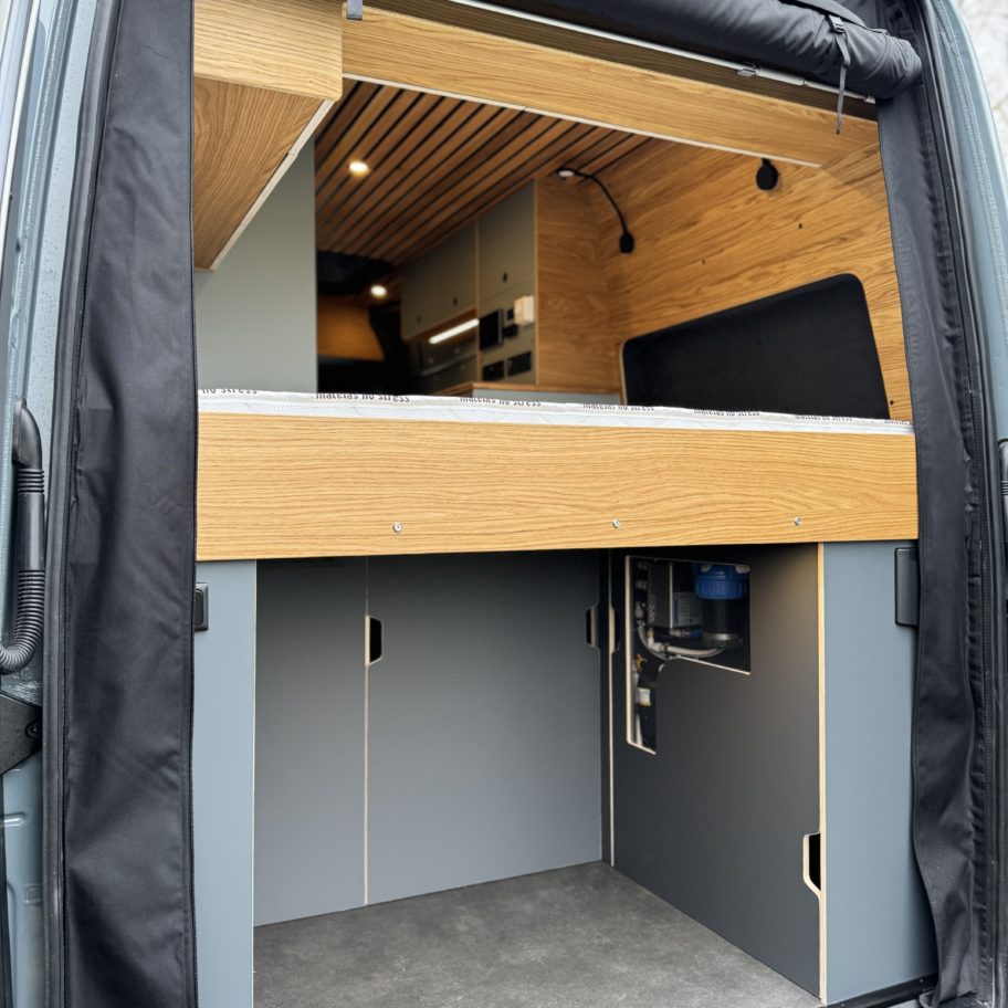 Espace de rangement arrière sous le lit d’un van Mercedes Sprinter aménagé en bois contreplaqué.