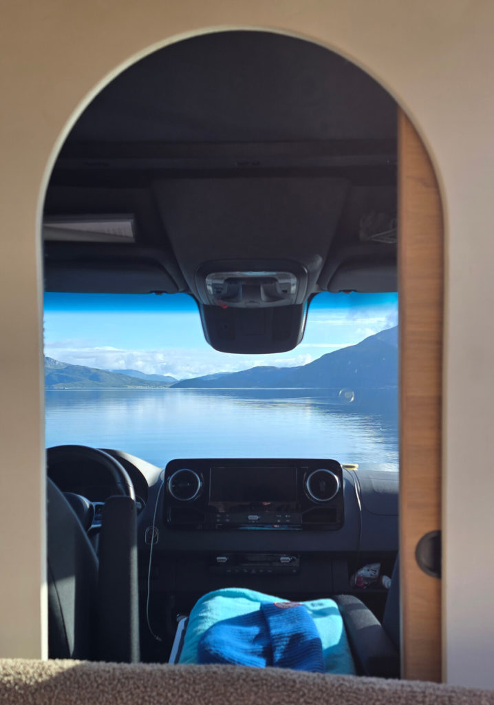Vue depuis l’intérieur d’un van aménagé à travers une porte arrondie, donnant sur un lac calme entouré de montagnes.