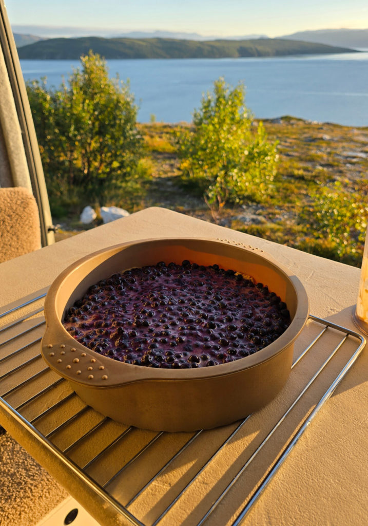 Gâteau aux myrtilles posé sur une grille, sur une table à l’intérieur d’un van, avec vue sur un lac et des collines en arrière-plan.