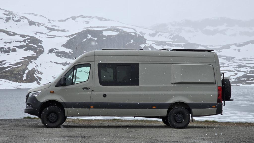 Van aménagé gris garé sur une route de montagne enneigée, avec des sommets couverts de neige en arrière-plan.