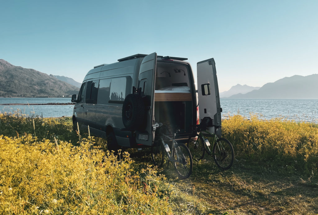 Van aménagé garé au bord d’un lac, portes arrière ouvertes, avec deux vélos posés contre le véhicule, dans un paysage naturel.